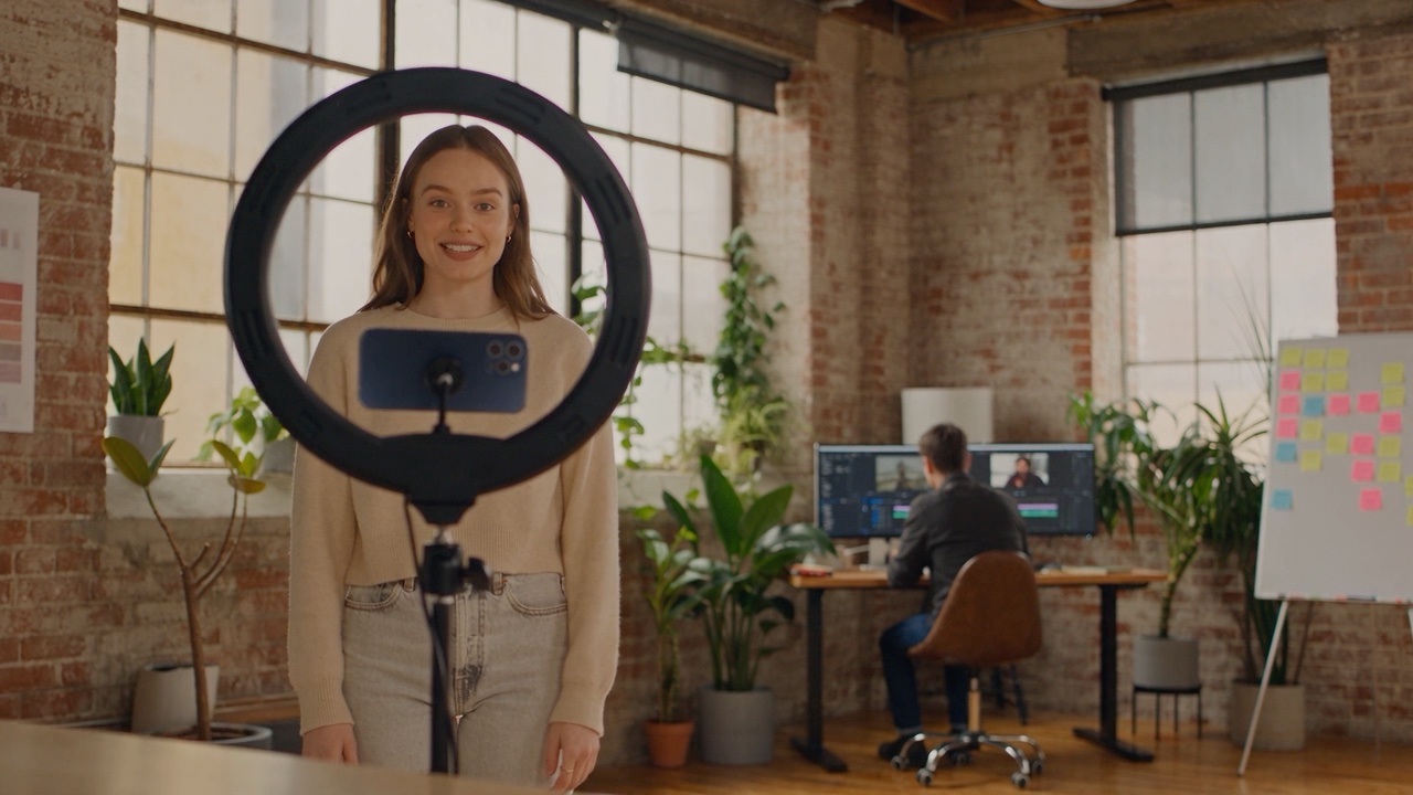A creator talking to a phone on a tripod inside a ring light, with an editor working at a timeline behind her